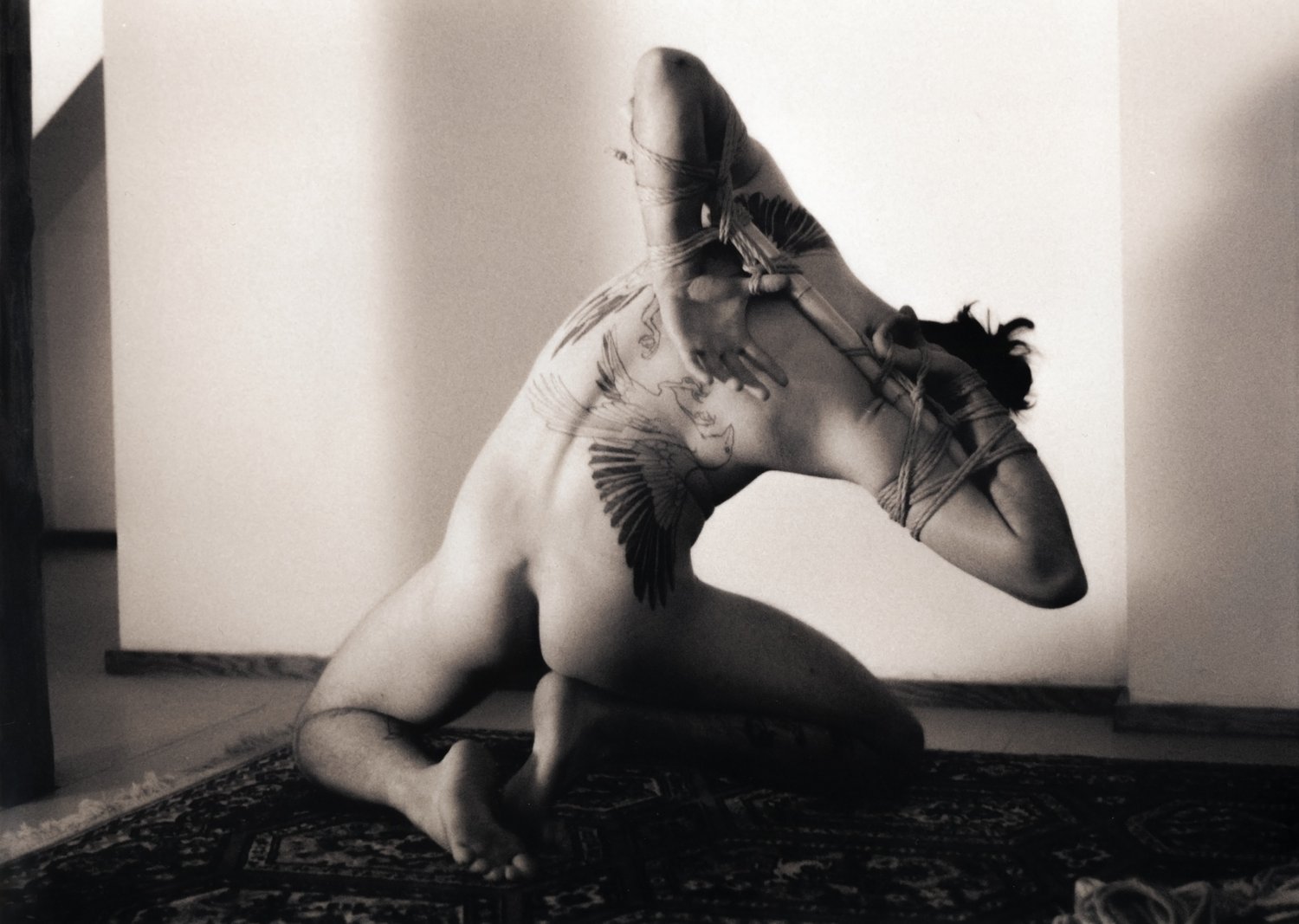 Naked tattooed person kneeling, bound with their arms in a teppou against bamboo bent over at their waist toward the right